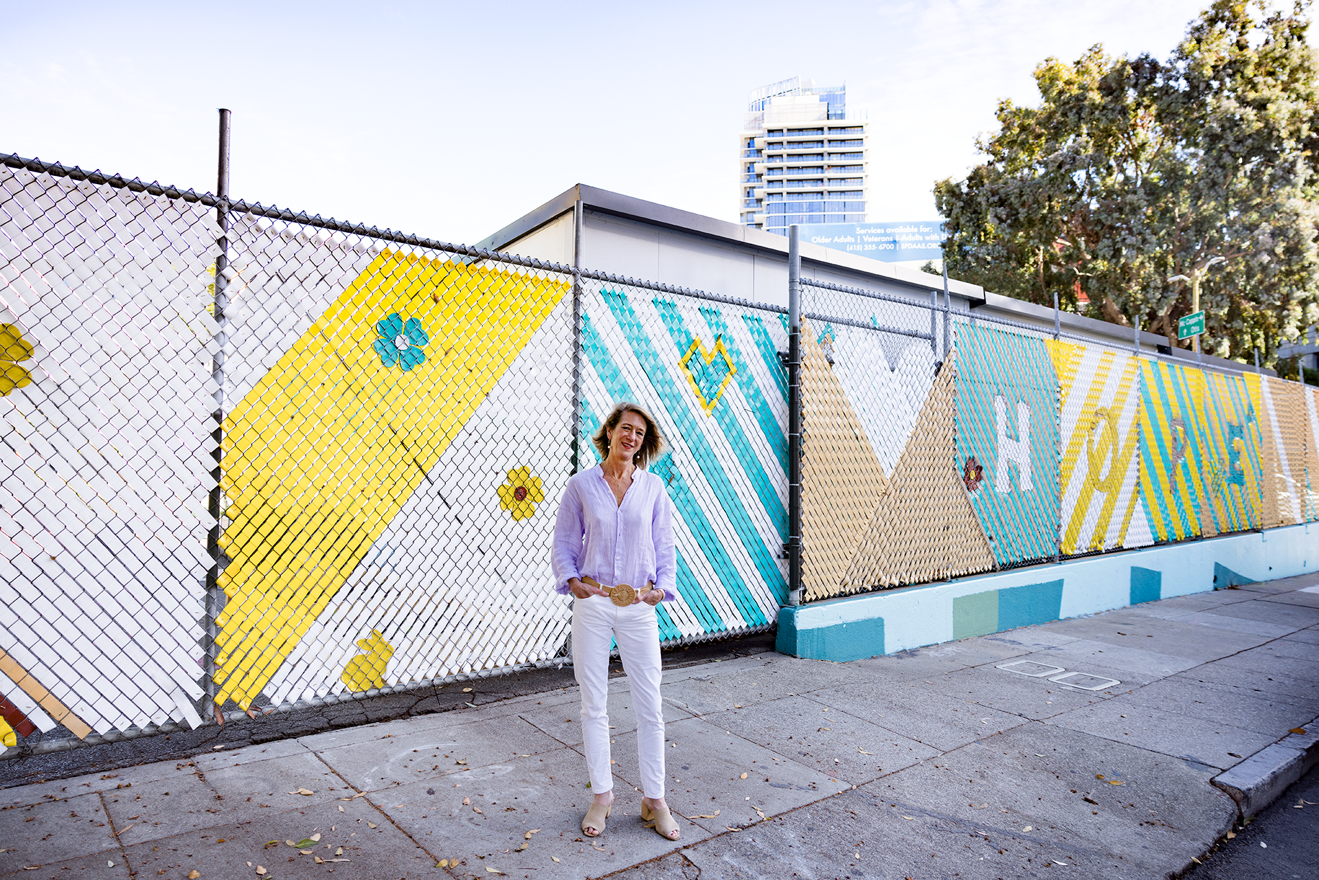 A photo of Elizabeth Funk standing in front of a large fence decorated with woven brightly colored ribbons with the word “hope” 