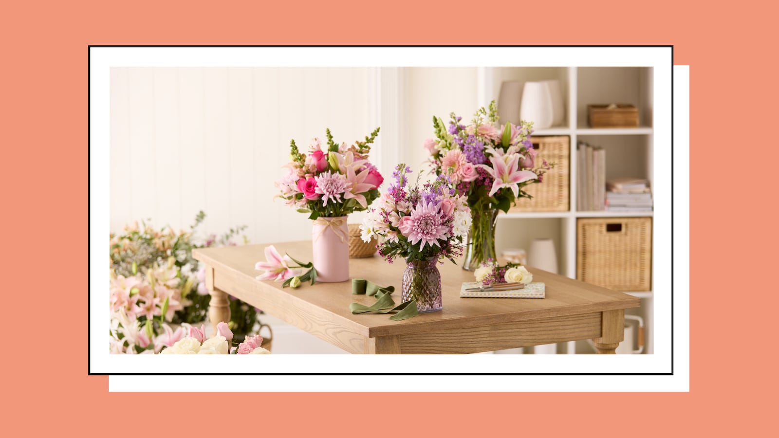 Three pink and purple spring flower bouquets arranged on a wooden table in a bright, airy room, surrounded by loose stems and greenery, with wicker storage shelving in the background.