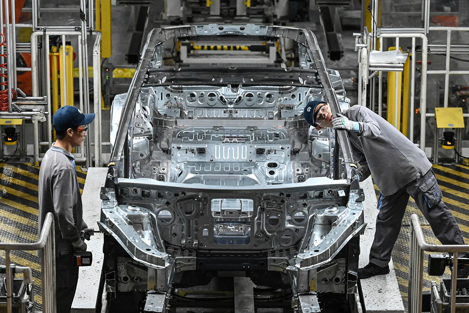 Two people on either side of a partially constructed electric vehicle on a production line. One person inspects the side of the car.