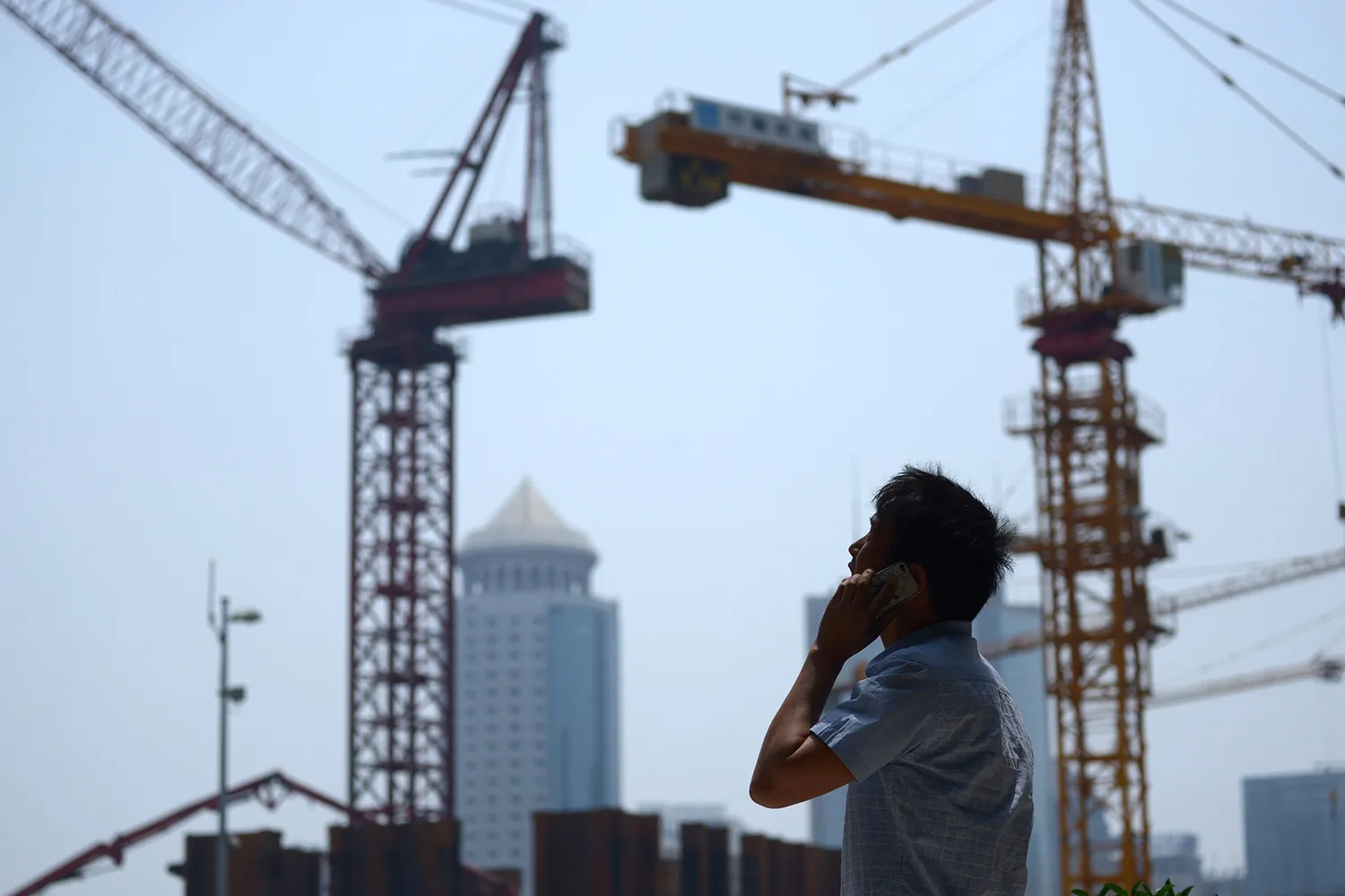 A man speaking on a cell phone looks up, facing a series of cranes and buildings in a city skyline.