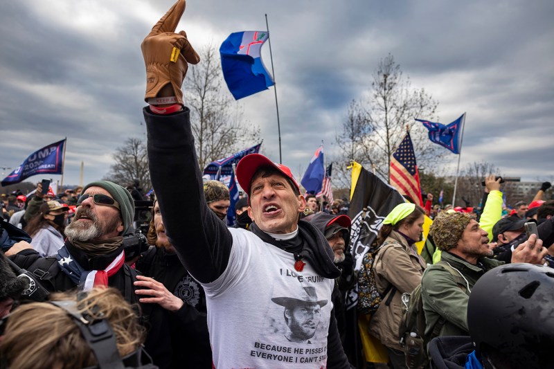 A crowd outdoors holding flags and signs. In the foreground, a man in a red baseball cap and white t-shirt raises a gloved hand with his index finger pointing upward while shouting.