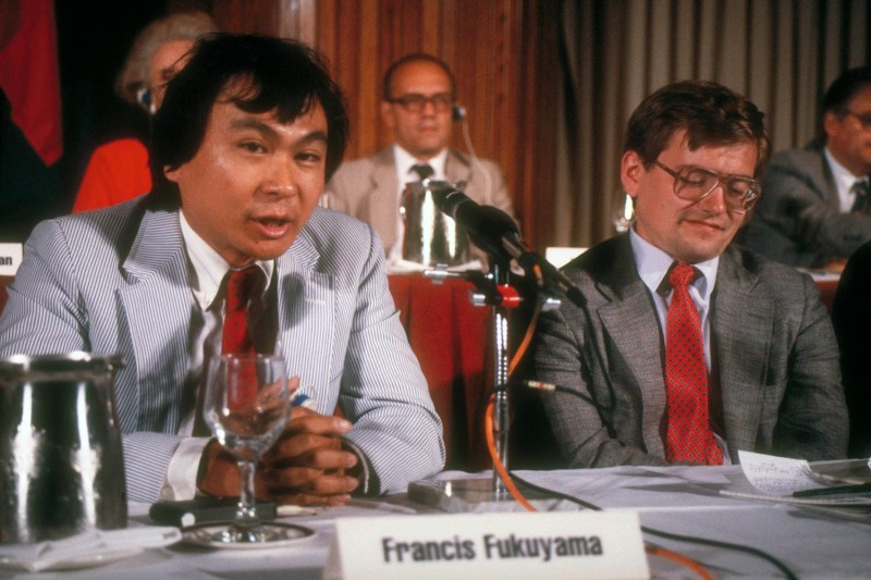 A man in a striped blazer and red tie speaks into a microphone at a panel table with a nameplate in front of him. Other panel participants sit on either side of him in front of a neutral backdrop.