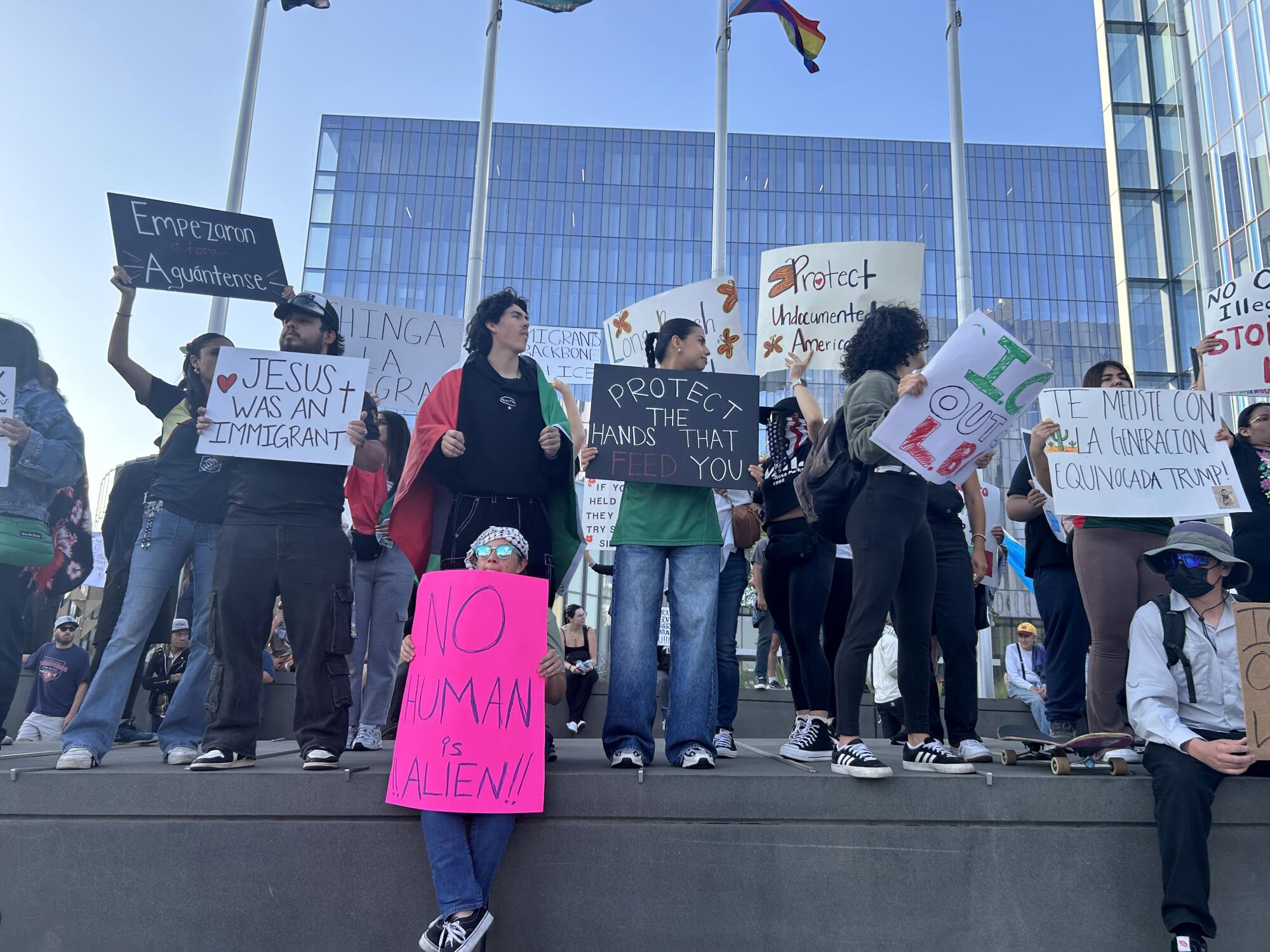 Protesters stand along a wall outside of Long Beach Civic Center holding signs.