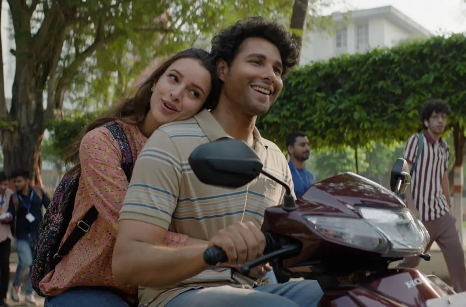 A man drives a scooter with a woman behind him, arms around his waist and head on his shoulder.