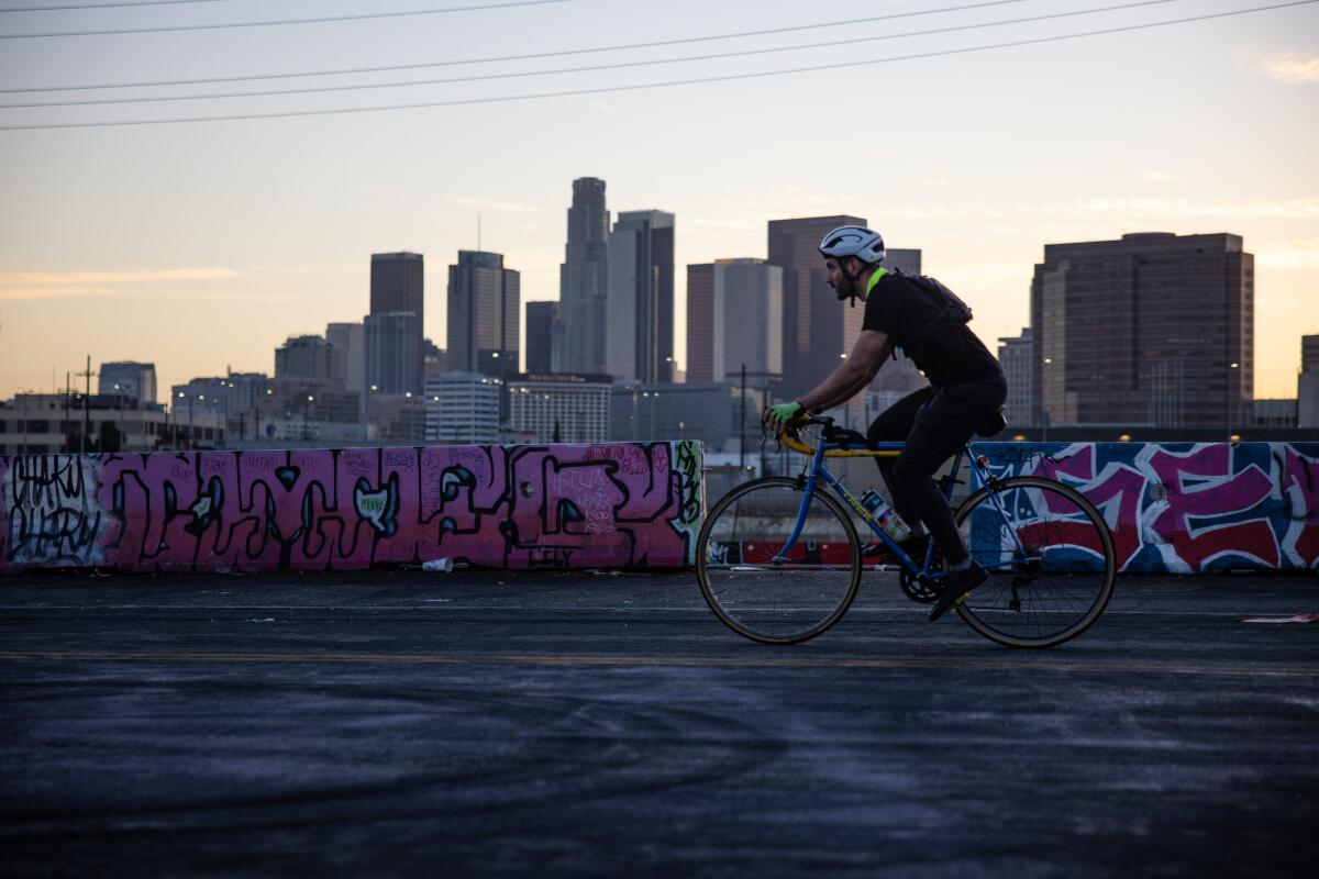 A cyclist rides a blue and yellow bike down a paved street with the downtown L.A. skyline in the distance.