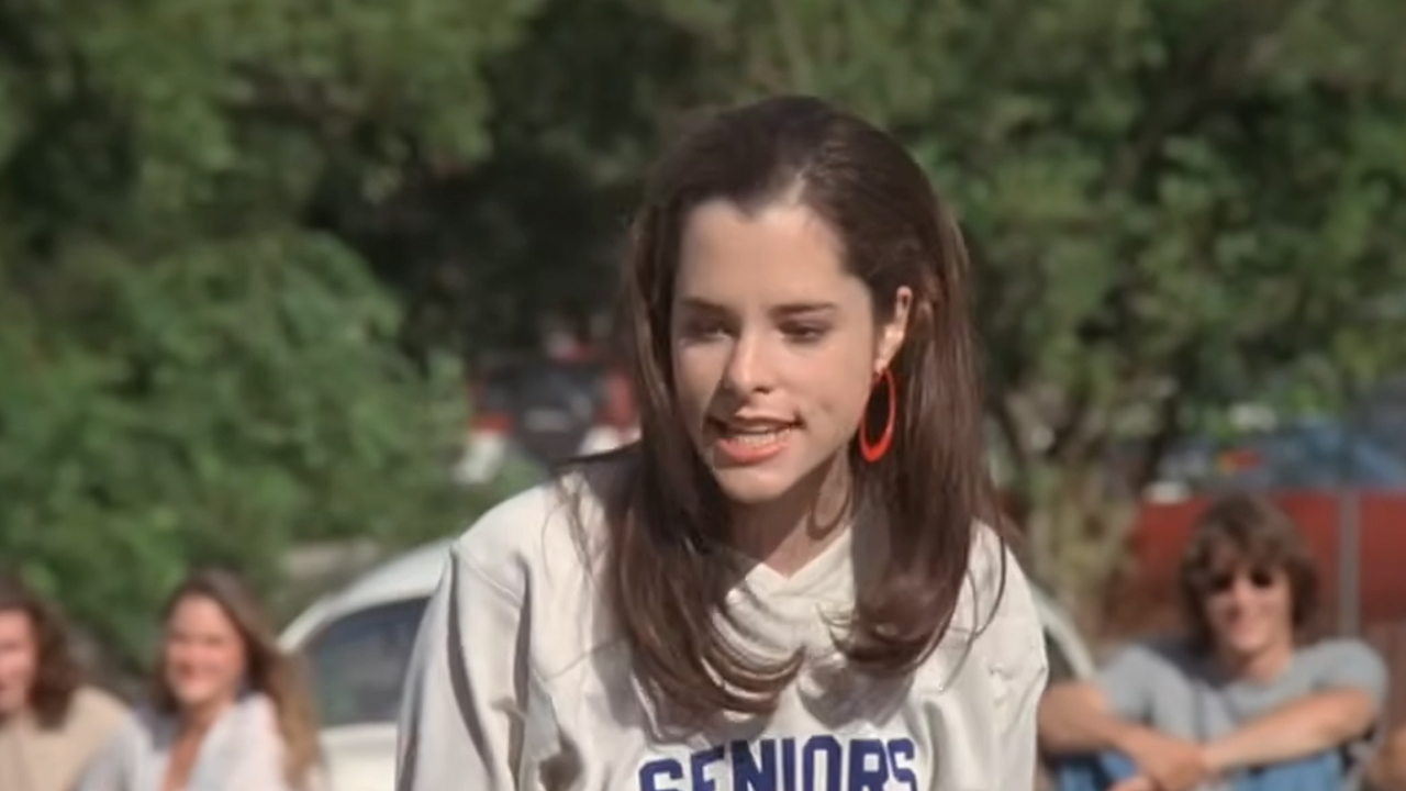 Parker Posey looking very upset and wearing a sweatshirt that says "seniors" on it in Dazed And Confused.