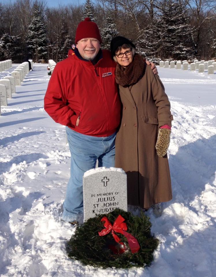 An older couple embraces and poses at the headstone of their family member Julius St. John Knudsen.