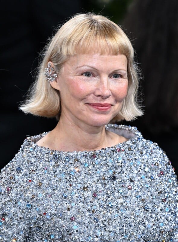 A woman with short blond hair and bangs wears a sparkling, jewel-covered outfit and a decorative ear cuff. She is smiling softly and standing against a dark, blurred background.