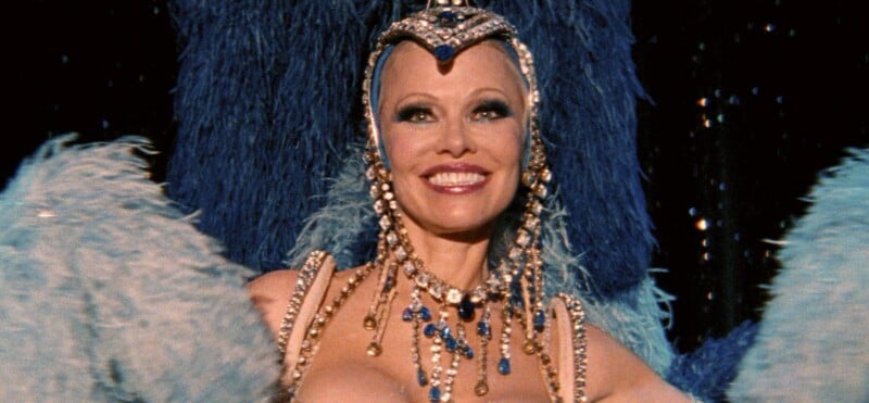 A smiling showgirl in an elaborate costume with blue and gold jewels, a beaded headdress, and large blue feathered wings performs on stage.