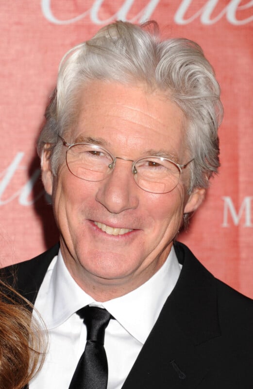 A mature man with gray hair, wearing glasses, a black suit, white shirt, and black tie, smiling in front of a red backdrop with white text.