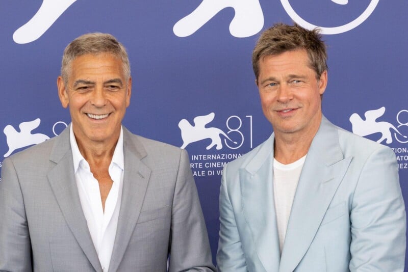 Two men in light-colored suits stand side by side, smiling, in front of a blue backdrop with white logos and text from the Venice International Film Festival.
