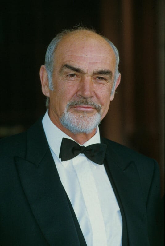 An older man with a receding hairline, white beard, and mustache is wearing a black tuxedo with a bow tie, standing and smiling slightly against a blurred dark background.