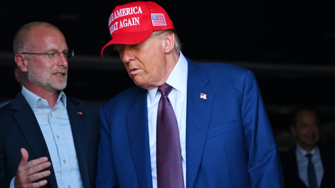 FCC Commissioner Brendan Carr standing next to and speaking to Donald Trump, who is wearing a 
