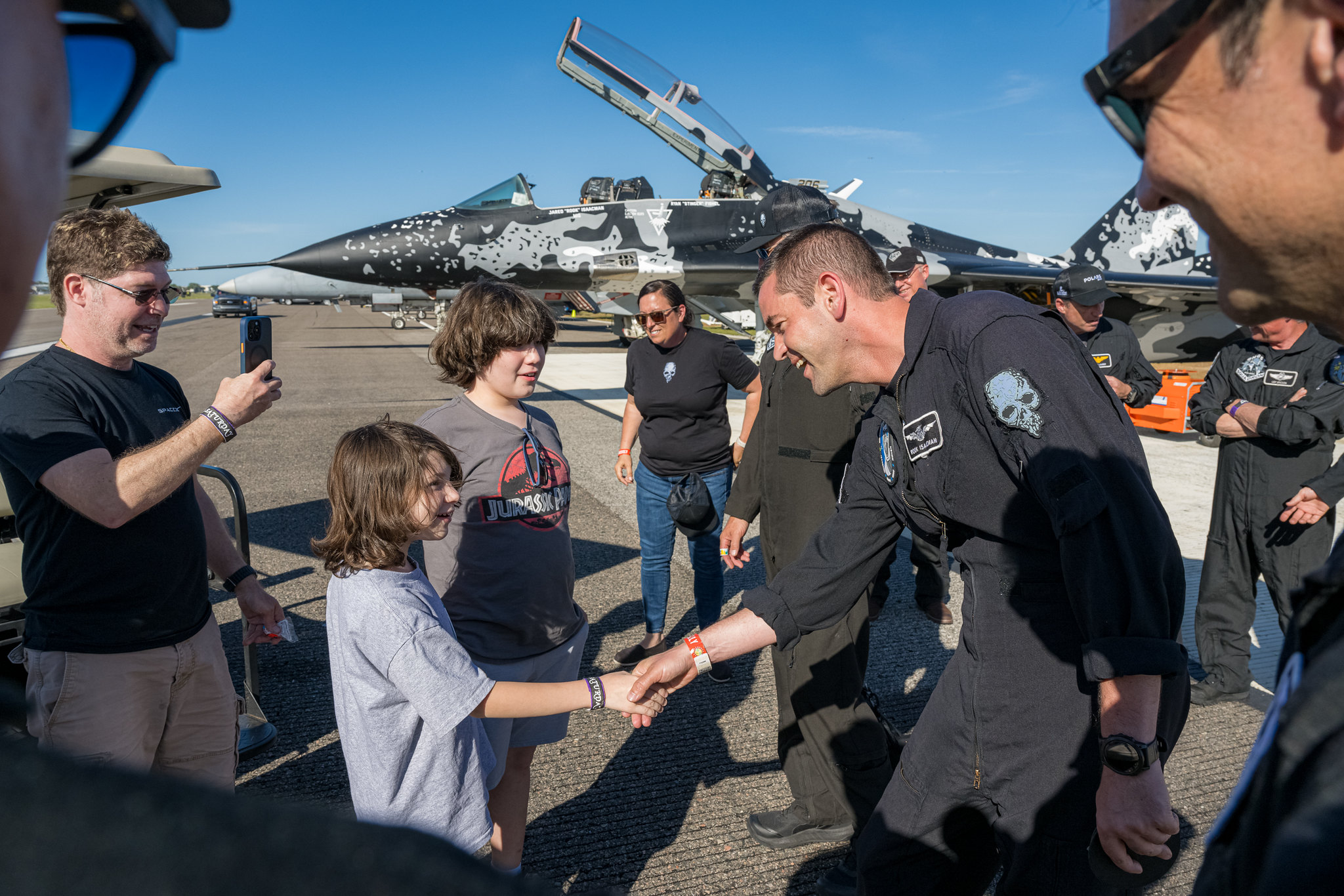 a smiling man in a blue flight suit greets people on a tarmac, with a fighter jet in the background