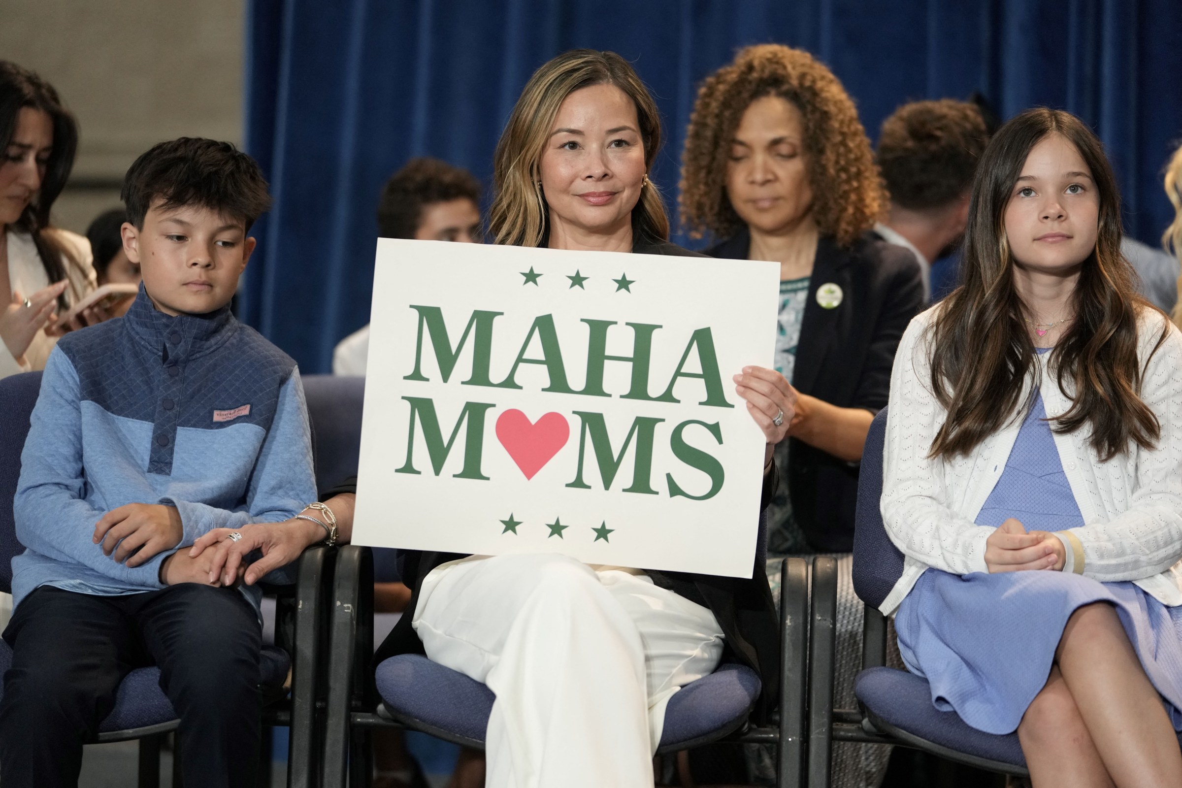 Mothers and children sit in rows of chairs, with one woman smiling and holding a sign reading “MAHA Moms.”