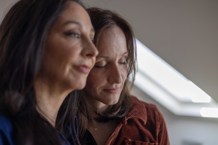 A close-up portrait of two women standing close together; the woman in the foreground looks off-camera with a soft expression, while the woman behind her closes her eyes gently with a peaceful smile.