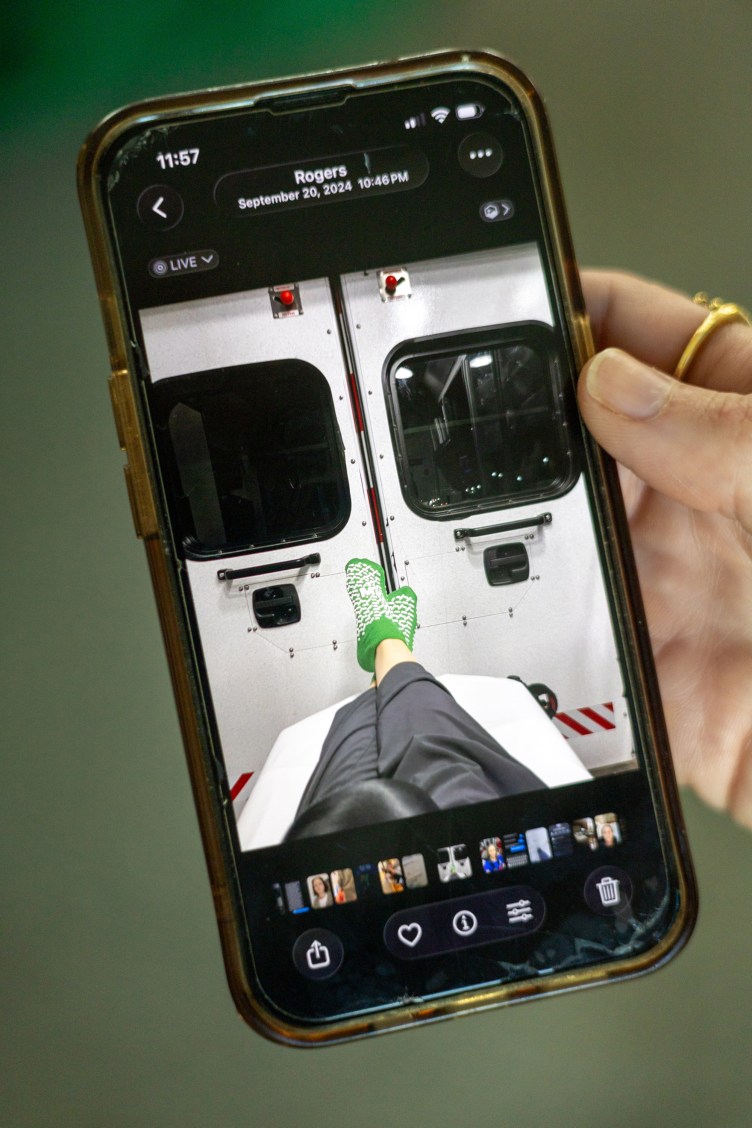 A hand holding a smartphone that displays a photo of a person's legs resting on a gurney inside an ambulance, wearing bright green hospital grip socks and facing the closed back doors.