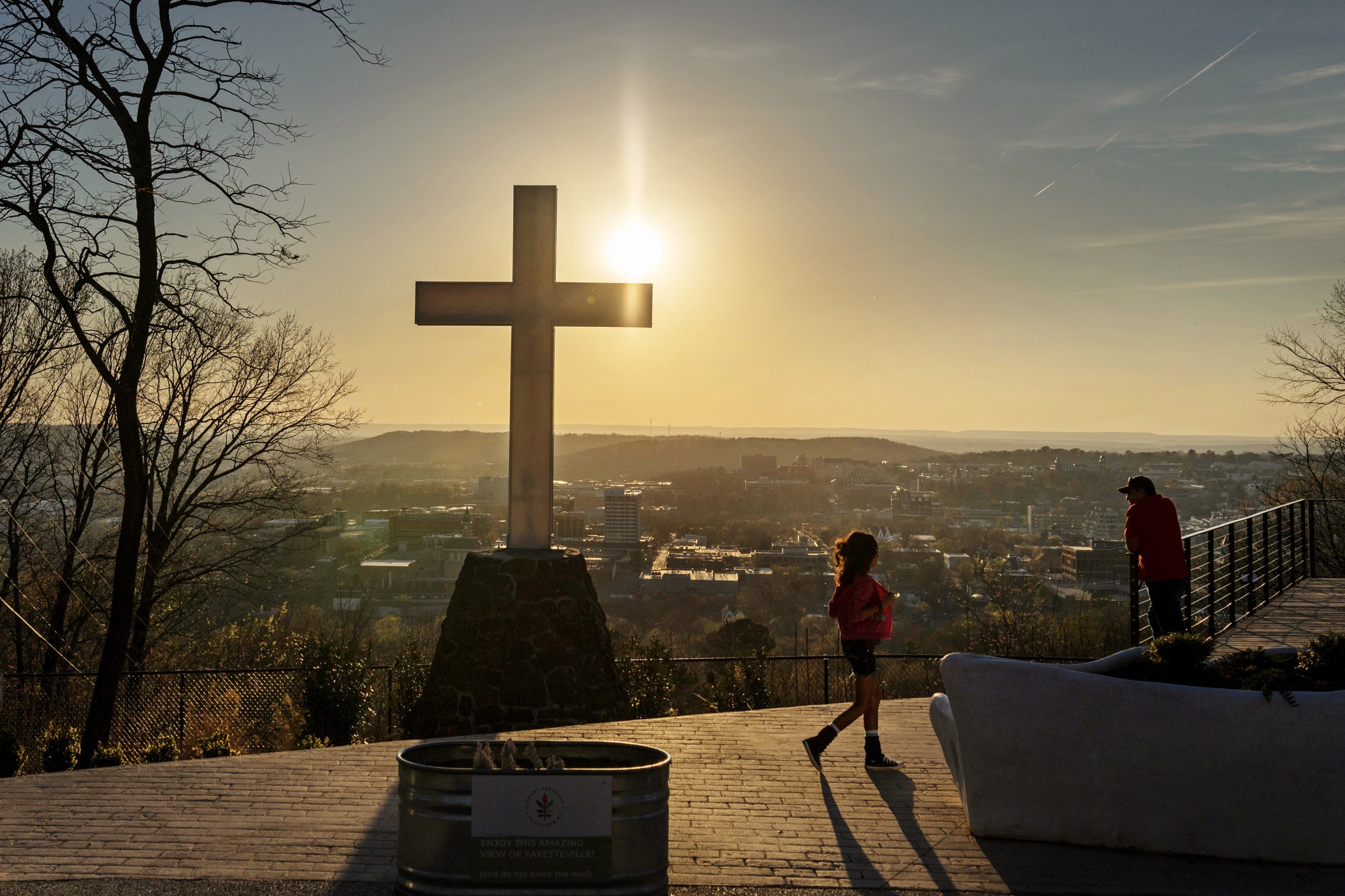 A large stone and metal cross stands on an overlook at sunset, framing a silhouette of a young girl walking and a person looking out over the hazy skyline of Fayetteville below.