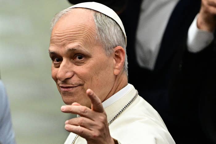 A man in religious attire gestures with a smile, appearing engaged and content