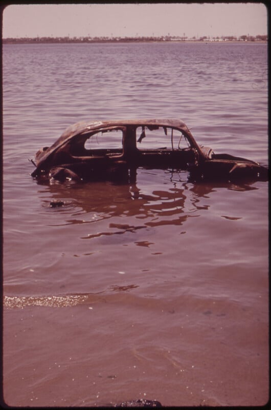 A rusted, partially submerged car sits in shallow water near a sandy shoreline, with distant trees and buildings visible across the water under a hazy sky.