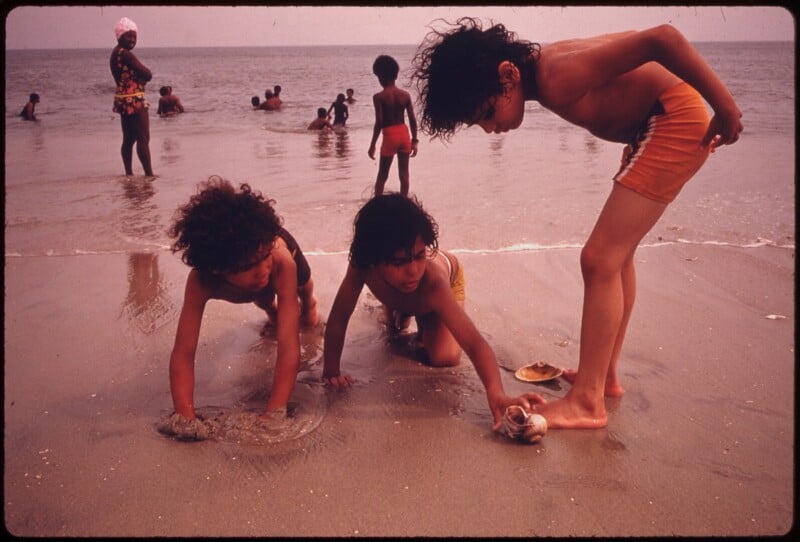 Three children play in the sand at the beach near the water, while other people swim and stand in the ocean in the background. The sky is overcast and the beach is wet.