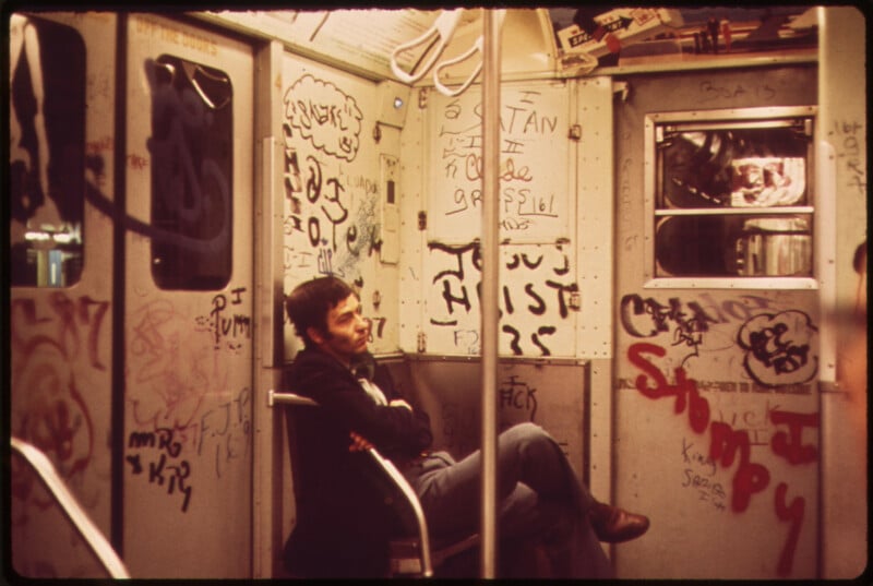 A man sits alone on a subway car covered in colorful graffiti. He is leaning back with his arms crossed, looking thoughtfully out of the frame. The walls are filled with various tags, drawings, and writing.