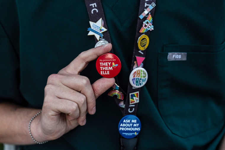 A hand holds a pin on a lanyard decorated with pronoun pins 